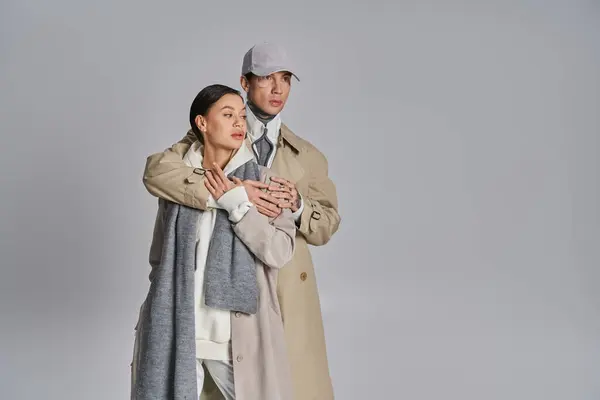 A fashionable young man and woman wearing trench coats stand side by side in a studio with a grey background. — Stock Photo