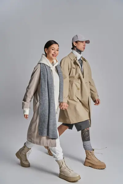 A stylish young couple, dressed in trench coats, walking together gracefully in a studio against a grey background. — Stock Photo