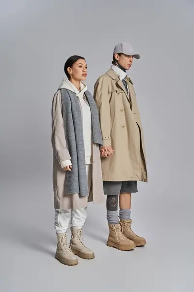 A young stylish couple in trench coats stand next to each other in a studio against a grey background. — Photo de stock