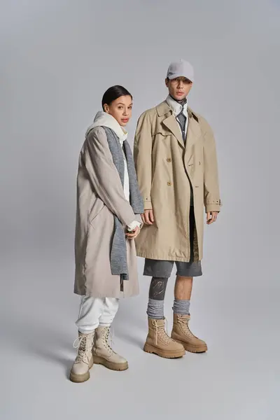 Young stylish couple in trench coats standing side by side in a studio, exuding urban elegance against a grey background. — Stock Photo