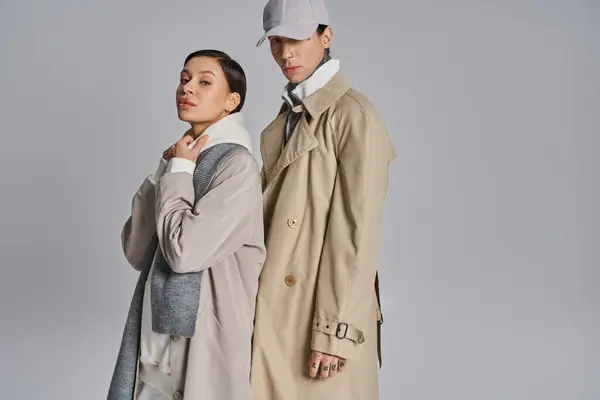 A young stylish couple in trench coats standing next to each other in a studio against a grey background. — Stock Photo