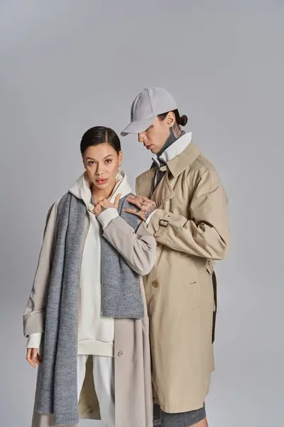 A young man and woman standing side by side, exuding style in trench coats against a grey studio background. — Stock Photo