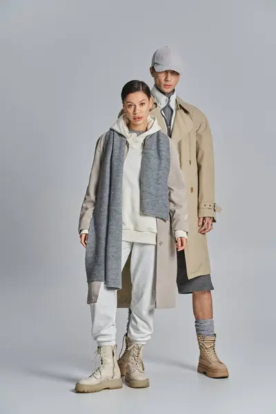 Ein junger Mann und eine junge Frau stehen nebeneinander in Trenchcoats, verströmen Stil und Eleganz in einem Studio mit grauem Hintergrund. — Stockfoto