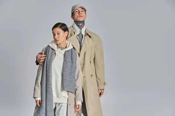 A young stylish couple wearing trench coats standing next to each other in a studio against a grey background. — Stock Photo