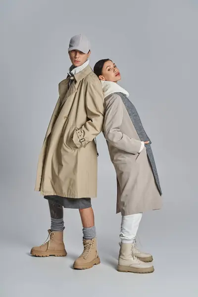 A young, stylish couple stands together in trench coats against a grey studio backdrop. — Stock Photo