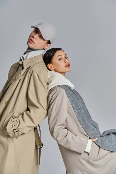 Un joven hombre y una mujer se paran elegantemente uno al lado del otro con elegantes abrigos de trinchera sobre un fondo gris de estudio. - foto de stock