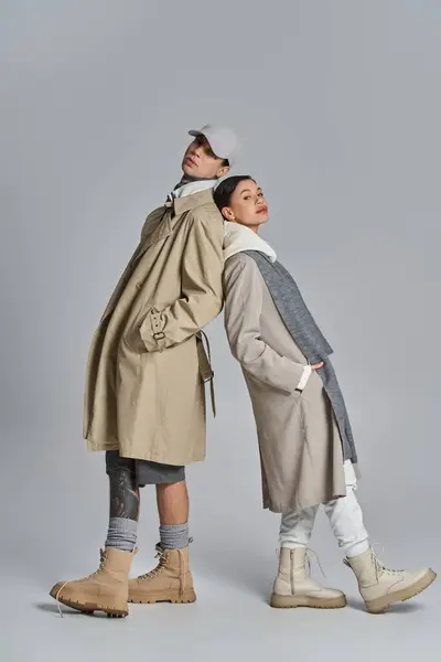 A stylish young man in a trench coat and hat standing next to another man in a similar outfit in a studio setting. — Stock Photo