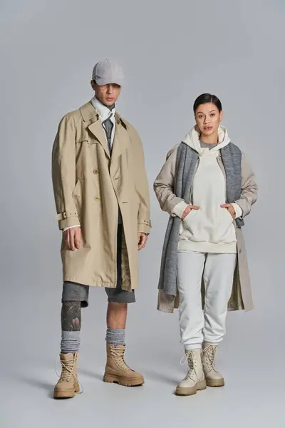 A young stylish couple in trench coats standing next to each other in a studio, against a grey background. — Stock Photo