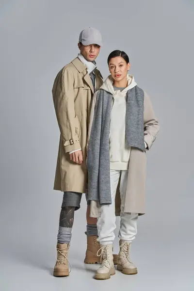 Ein junger Mann und eine junge Frau in schicken Trenchcoats stehen nebeneinander in einem Atelier vor grauem Hintergrund. — Stockfoto