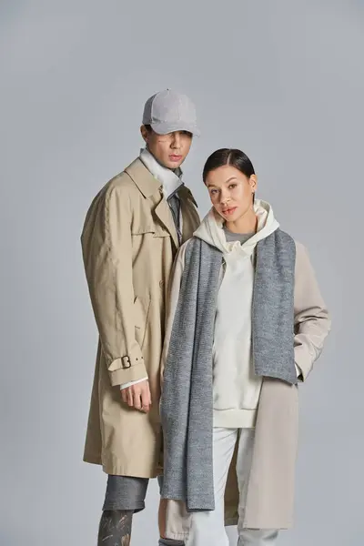 A young stylish couple stands side by side in trench coats in a studio against a grey background. — Stock Photo