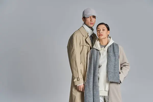 Una pareja joven y elegante se encuentra lado a lado en abrigos de trinchera contra un fondo gris del estudio. - foto de stock