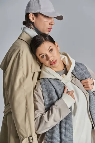 Ein stilvoller junger Mann und eine junge Frau stehen Seite an Seite in Trenchcoats vor grauer Studiokulisse. — Stockfoto