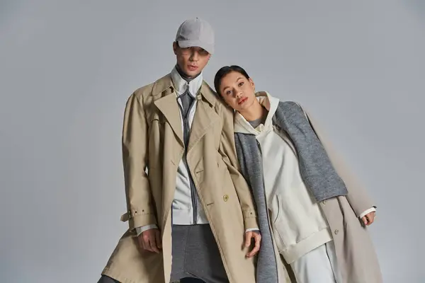 Ein junges, stylisches Paar steht selbstbewusst Seite an Seite in Trenchcoats vor grauem Hintergrund. — Stockfoto