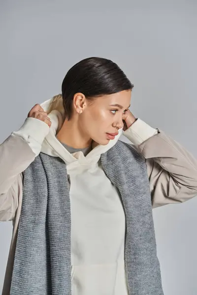 A young woman clad in a white top gracefully drapes a gray scarf, exuding sophistication in a studio against a grey background. — Stock Photo