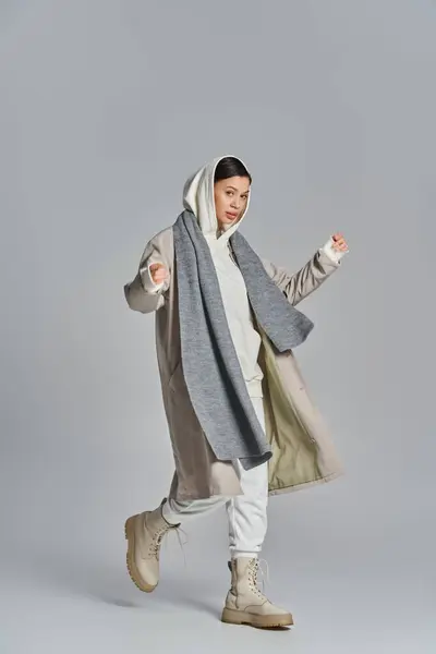 Uma mulher está confiante em um casaco cinza e branco e calças brancas, exalando estilo e sofisticação em um pano de fundo cinza. — Fotografia de Stock