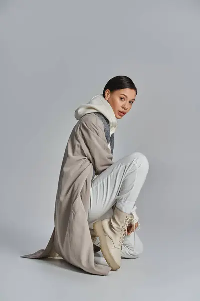 Eine junge, stylische Frau sitzt auf dem Boden, modisch im Trenchcoat, vor grauem Studiohintergrund. — Stockfoto