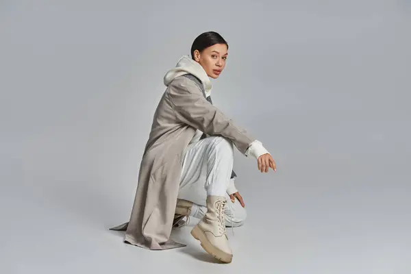 A young stylish woman in a trench coat sitting gracefully in a studio against a grey background. — Stock Photo