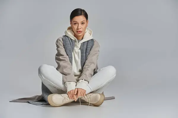 A young stylish woman in a trench coat sits peacefully on the floor, legs crossed, lost in thought in a studio setting with a grey background. — Stock Photo