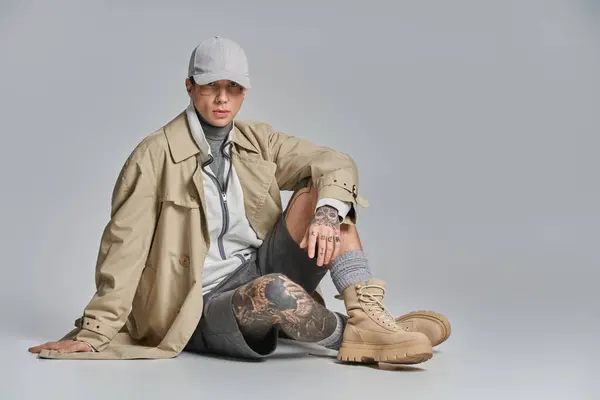 Ein junger tätowierter Mann im Trenchcoat sitzt mit überkreuzten Beinen und strahlt auf grauem Studiohintergrund Nachdenklichkeit aus.. — Stockfoto