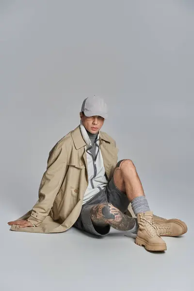 A young, tattooed man in a trench coat sitting on the floor with his legs crossed in a studio on a grey background. - foto de stock