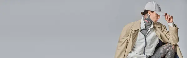 A young tattooed man exudes mystery and style in a trench coat and hat, standing confidently in a studio against a grey backdrop. — Stock Photo