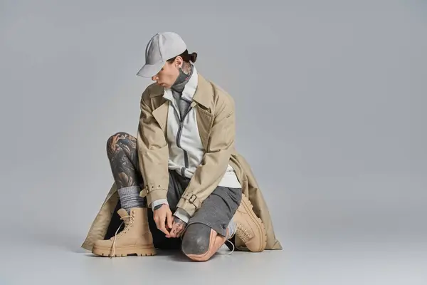 Ein junger Mann mit Tätowierungen sitzt mit Hut und Trenchcoat auf dem Boden vor grauem Studiohintergrund.. — Stockfoto