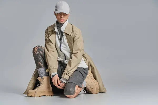 Ein junger, tätowierter Mann sitzt auf dem Boden, feierlich in einen Trenchcoat gehüllt vor grauer Studiokulisse. — Stockfoto