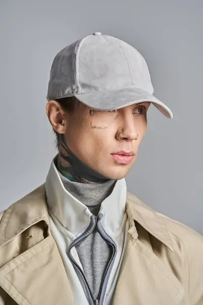 Un giovane uomo tatuato trasuda freschezza in un elegante trench, completo di cappello alla moda, sullo sfondo di uno studio grigio. — Foto stock