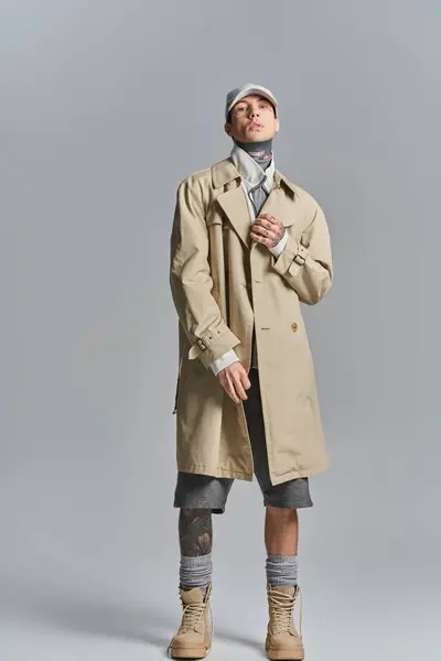 Um jovem, tatuado, coloca confiantemente em um casaco de trincheira em um estúdio contra um fundo cinza. — Fotografia de Stock