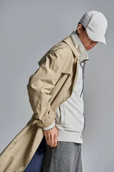 A stylish young man with tattoos wears a trench coat and hat, exuding an air of mystery in a studio setting against a grey background. — Stock Photo