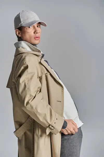 A stylish man with a striking hat and elegant trench coat poses confidently in a studio setting against a grey background. — Stock Photo