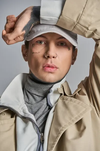 A young tattooed man in a trench coat stands confidently, wearing a stylish hat on his head in a studio setting against a grey background. — Stock Photo