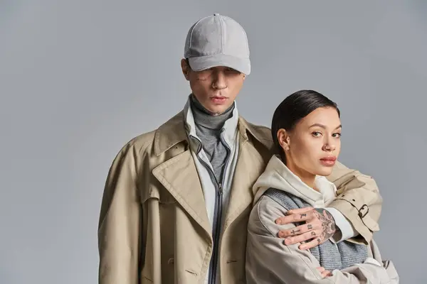 Un joven hombre y una mujer se paran uno al lado del otro con abrigos de trinchera, exudando estilo y gracia en un estudio sobre un fondo gris. — Stock Photo