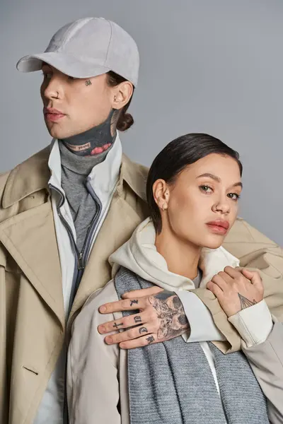 Un joven hombre y una mujer se colocan con estilo juntos en abrigos de trinchera sobre un fondo gris estudio. - foto de stock