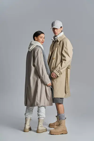 Ein junger Mann und eine junge Frau stehen stilvoll nebeneinander in Trenchcoats in einem Atelier vor grauem Hintergrund. — Stockfoto