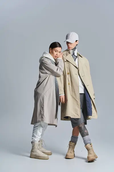 Um jovem casal elegante está lado a lado em casacos de trincheira, exalando sofisticação e elegância em um estúdio contra um fundo cinza. — Fotografia de Stock