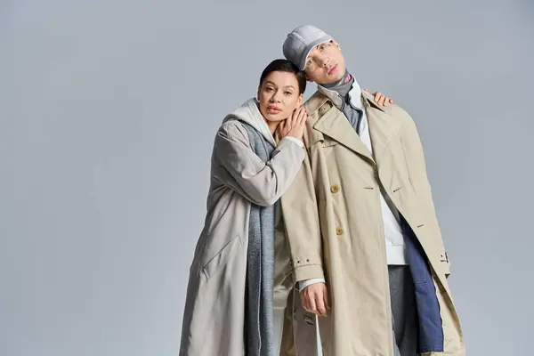 A young, stylish couple stands side by side, exuding elegance in trench coats, in a studio against a grey background. — Stock Photo