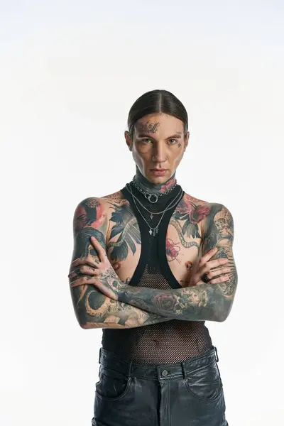 Um jovem elegante com tatuagens fica confiante, braços cruzados, em um estúdio contra um fundo cinza. — Fotografia de Stock