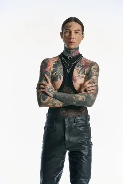 Стильний молодий чоловік з татуюваннями стоїть впевнено, перетинаючи руки в студії на сірому фоні. — стокове фото
