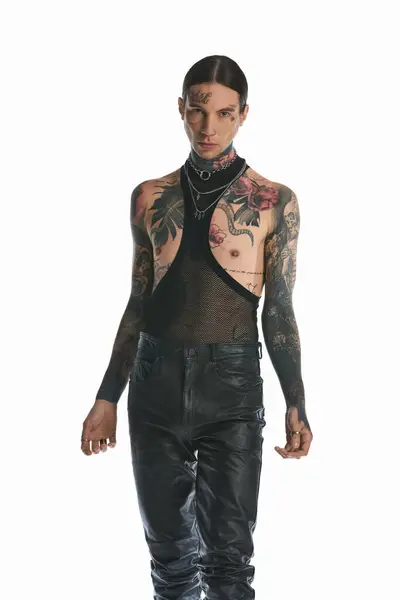 Un giovane uomo tatuato sul suo corpo, vestito di pantaloni neri, posa in uno studio su uno sfondo grigio. — Foto stock