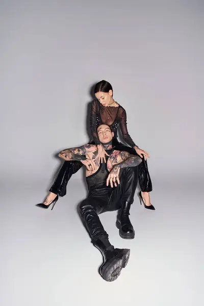 Stilvolles junges Paar, Mann mit Tätowierungen, sitzt auf dem Rücken von Frauen in einem Studio vor grauem Hintergrund. — Stockfoto