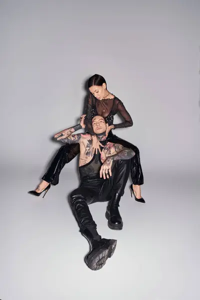 Молодая стильная женщина сидит на вершине мужчины в студии, оба покрыты татуировками, на сером фоне. — стоковое фото
