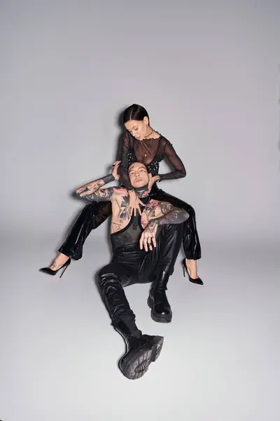 A young stylish woman is seated on top of a man in a studio, both covered in tattoos, against a grey background. — Stock Photo
