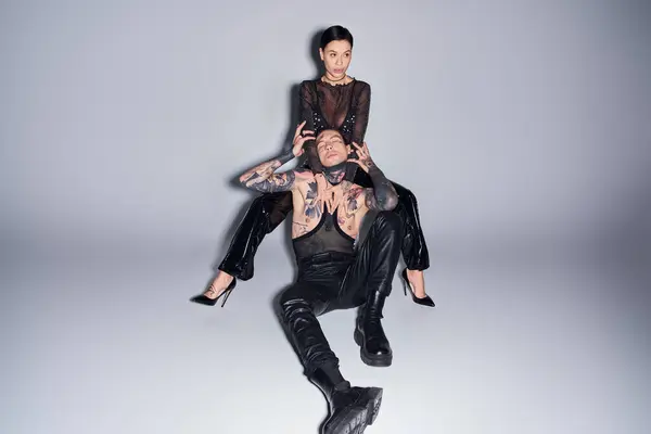 A stylish, tattooed couple in a studio setting, the woman sitting on top of the man against a grey background. — Stock Photo