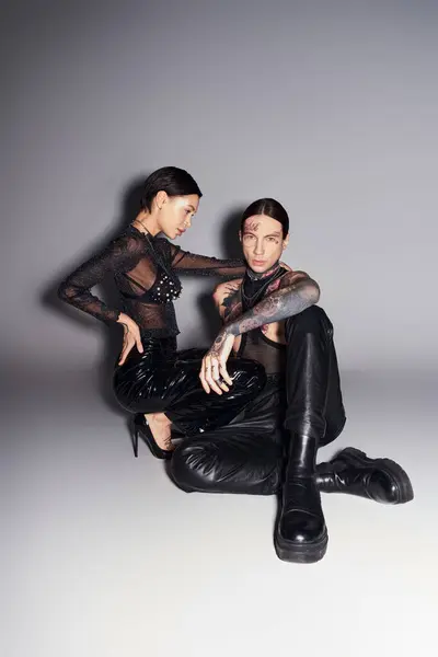 Deux jeunes femmes élégantes avec des tatouages assis ensemble dans un studio sur un fond gris. — Photo de stock