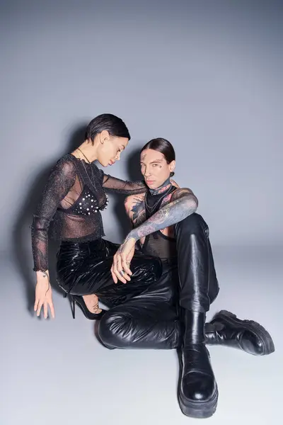 Un jeune homme et une jeune femme élégants avec des tatouages assis côte à côte dans un studio sur un fond gris. — Photo de stock