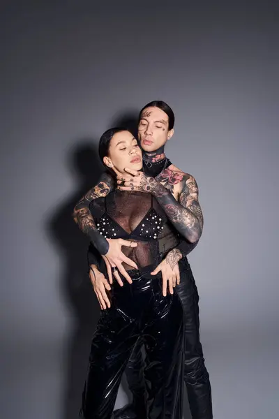 A young, stylish couple with tattoos dressed in matching black leather outfits pose in a studio against a grey background. — Stock Photo