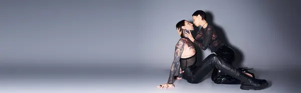 Стильная, татуированная молодая женщина изящно сидит на земле рядом с мужчиной в студии на сером фоне. — стоковое фото