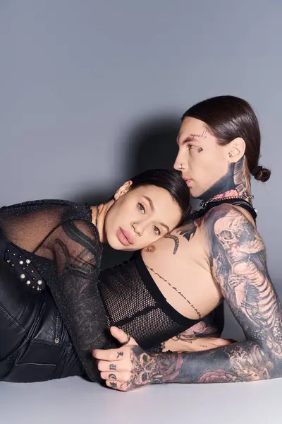 A young stylish couple, adorned with tattoos, lying down together in a loving embrace on a grey studio background. — Stock Photo