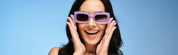 Une femme élégante avec des lunettes de soleil violettes, mains sur le visage, respirant la confiance dans un tournage en studio d'été sur un fond bleu. — Photo de stock